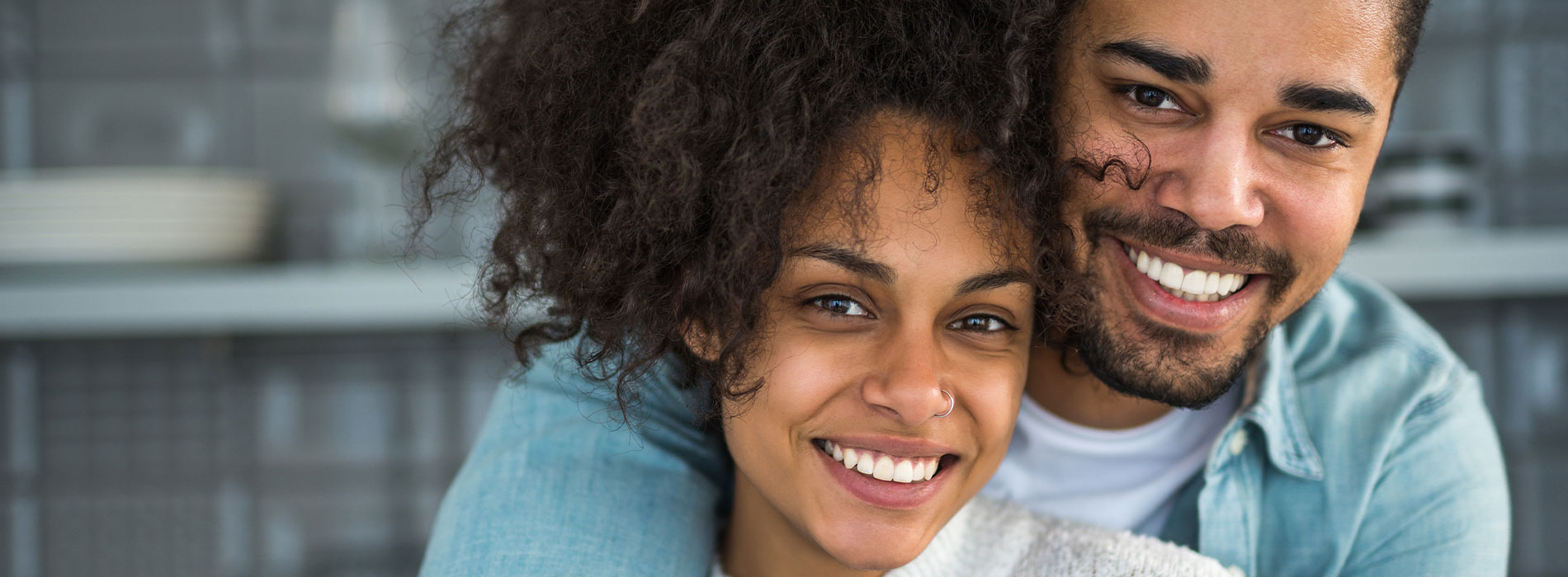A man and woman are smiling at each other, with the man's arm wrapped around the woman, set against a blurred kitchen background.