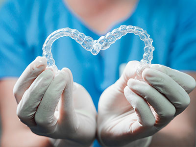 A person's hands cradling a clear plastic heart-shaped object, which appears to be a dental retainer, against a blue background with medical gloves visible.