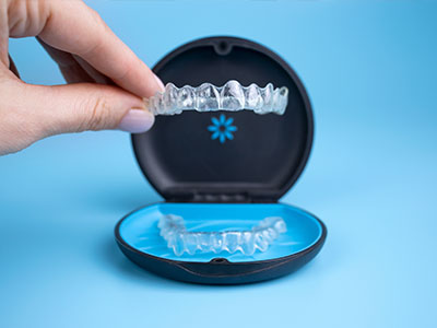 The image shows a person's hand holding a clear plastic tray containing a set of transparent dental aligners, with an open blue case displaying one of these aligners in front.