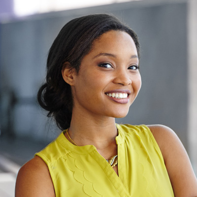 The image shows a smiling woman with dark hair wearing a yellow top, standing against a blurred background, possibly posing for a portrait.