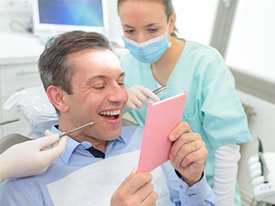The image shows a man sitting in a dental chair with a smile on his face, holding up a pink card with a surprised expression while looking at it. In the background, there's a female dental professional wearing gloves and a mask, standing behind him, smiling as well.