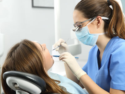 A dental hygienist performing a cleaning procedure on a patient's teeth while wearing protective gloves and a mask, with a woman seated in the dental chair receiving the service.