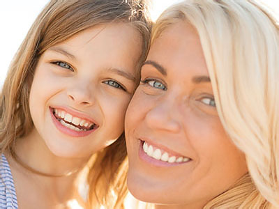 A woman and young girl are smiling at the camera; they appear to be enjoying a sunny day outdoors.