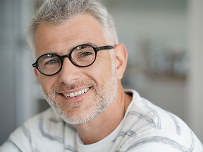 The image features a smiling man with gray hair and glasses, wearing a white shirt with a patterned collar, looking directly at the camera.
